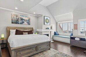 A bedroom with a large bed, a fireplace, a window with blinds, and artwork on the walls. - Pergola Terrace + Fireplace + Pool + Gym | Salt Creek + Harbor | Terracotta (Dana Point)