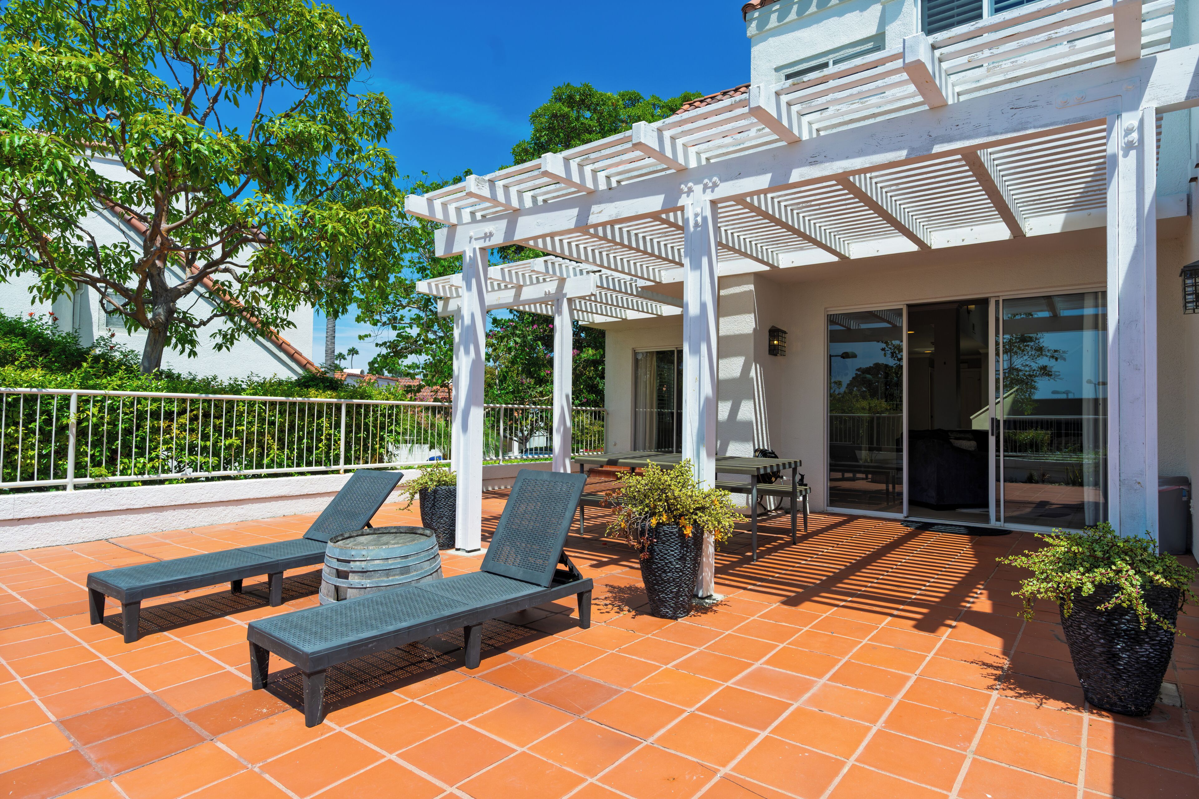 A covered outdoor seating area with two lounge chairs and a table, a pergola with white beams, and a tiled patio.