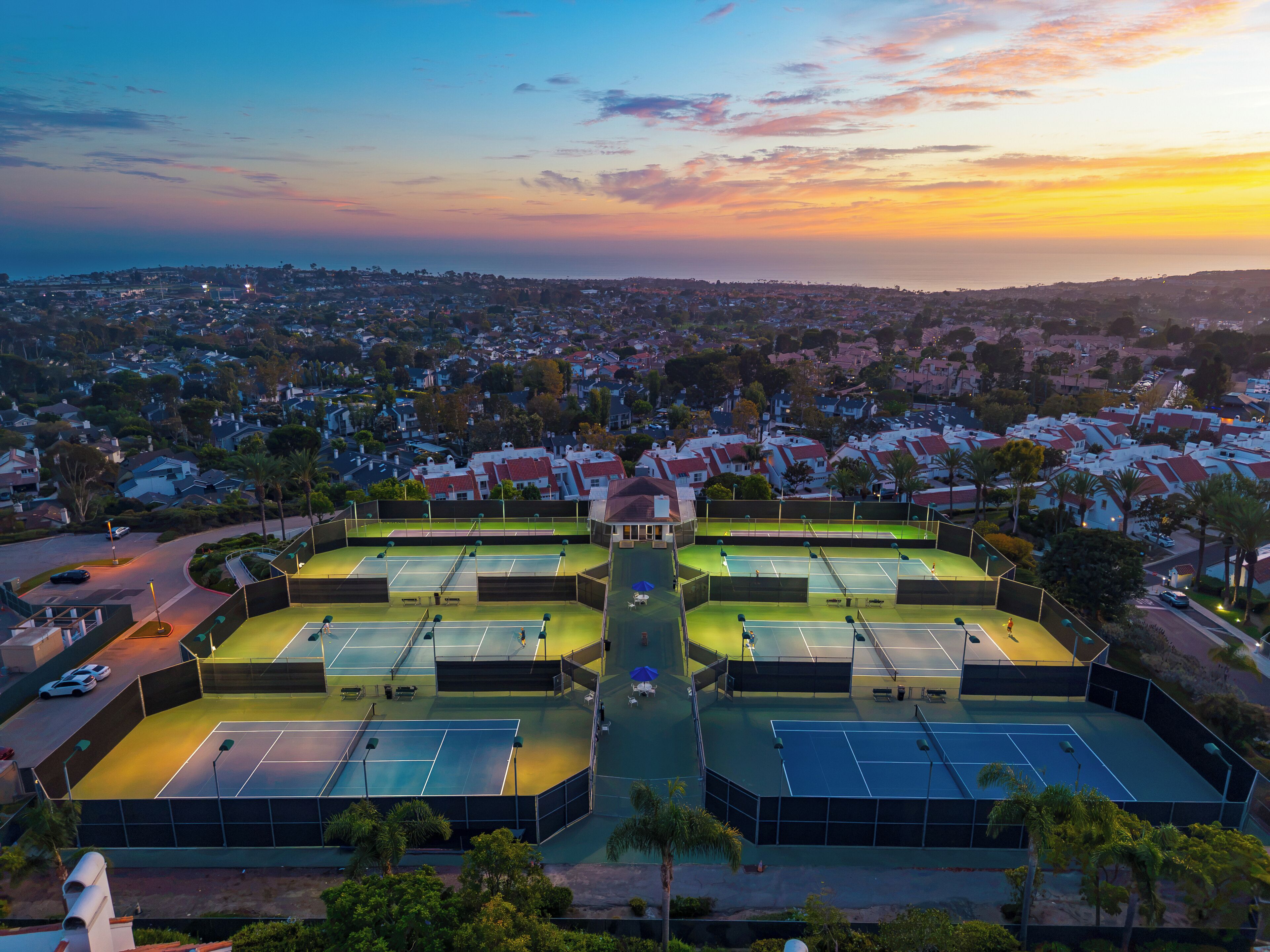 A tennis court complex with multiple courts, surrounded by residential buildings and palm trees.