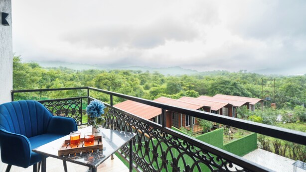 Un balcon avec une chaise bleue, une petite table avec des boissons, et une vue sur un paysage verdoyant avec des bâtiments visibles au loin.