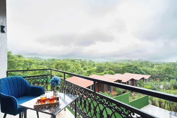 A balcony with a blue chair, a small table with drinks, and a view of a green landscape with buildings in the distance.