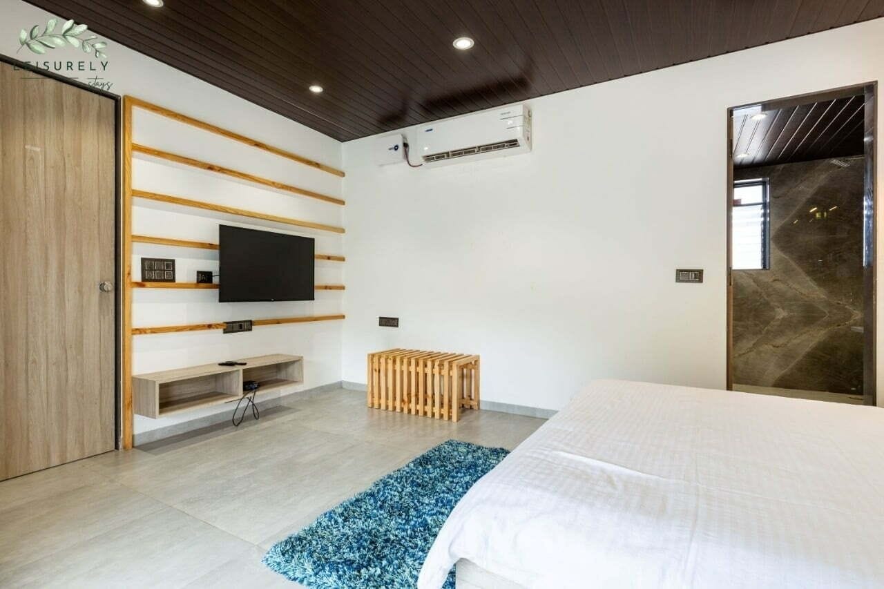 Un dormitori modern amb un llit, una televisió penjada a la paret, una banca de fusta i una taula petita.