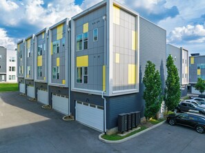 A multi-story building with a modern design, featuring a combination of grey and yellow panels, large windows, and a parking area with several cars. - Open-Concept Design + Game Stash | Mins to Five Points | Harmony House (Nashville)