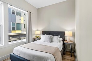 Una habitació d'hotel amb un llit gran, dues taules de nit amb làmpades, una finestra amb cortines i vista a l'edifici exterior.