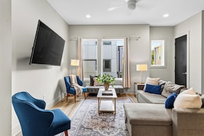 A modern living room with a flat-screen TV mounted on the wall, a sofa, a chair, a coffee table, and a window with curtains. - Open-Concept Design + Game Stash | Mins to Five Points | Harmony House (Nashville)