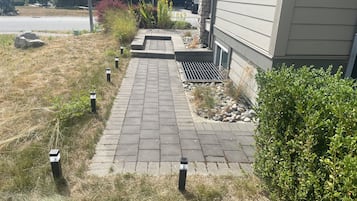 Un chemin pavé mène à une maison dont les fondations sont en pierre, et qui est bordée d’un petit jardin agrémenté de rochers et d’arbustes.