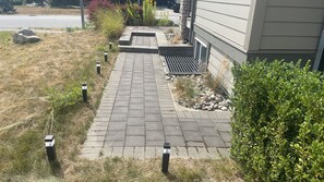 Un sendero empedrado que conduce a una casa con base de piedra y un pequeño jardín con rocas y arbustos.