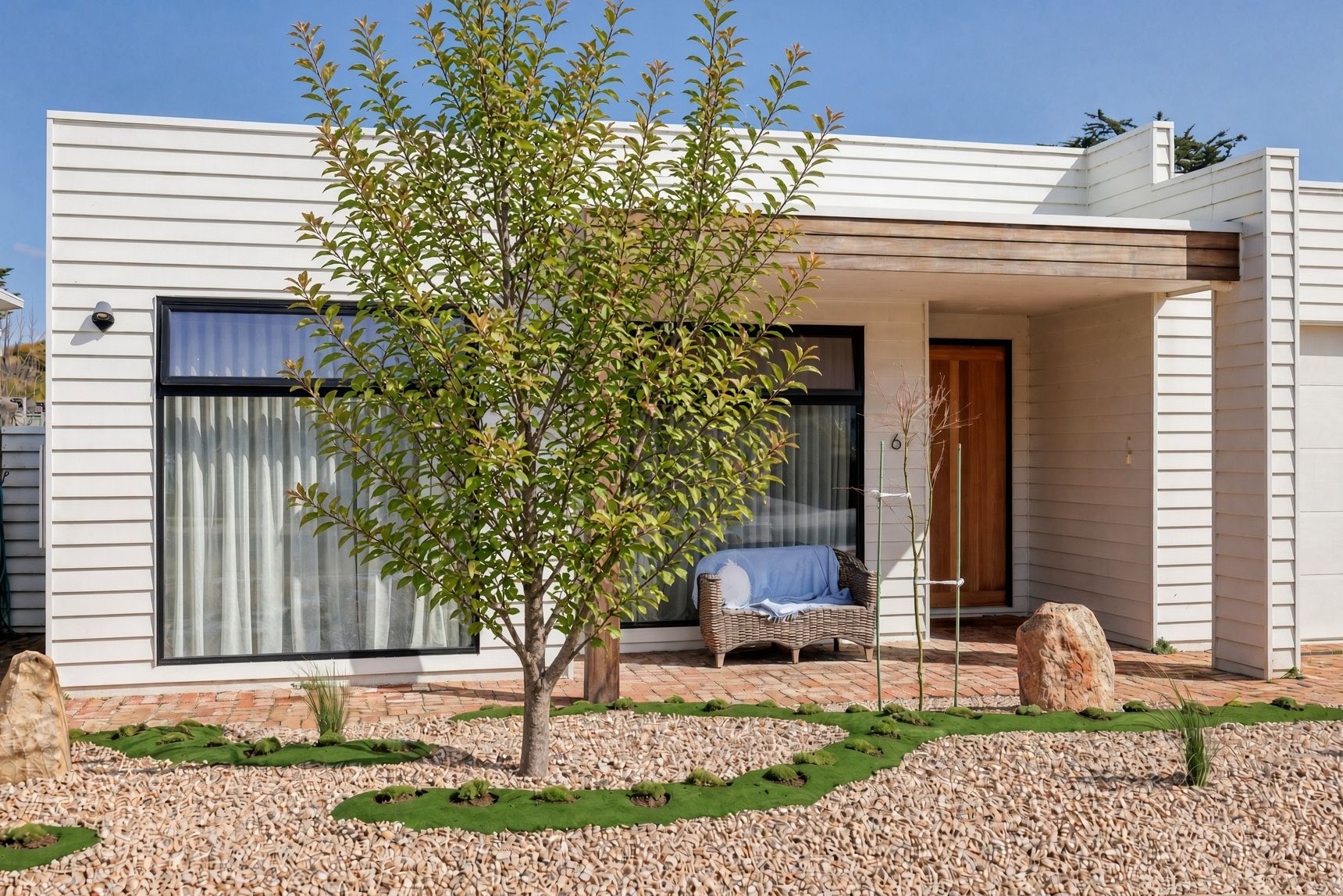 Une maison moderne dotée d’un porche abrité, d’un petit arbre et d’un canapé en osier.