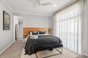 Ein Schlafzimmer mit einem Bett, einem Deckenventilator, einem Fenster mit transparenten Vorhängen und einem zur Toilette führenden Gang.