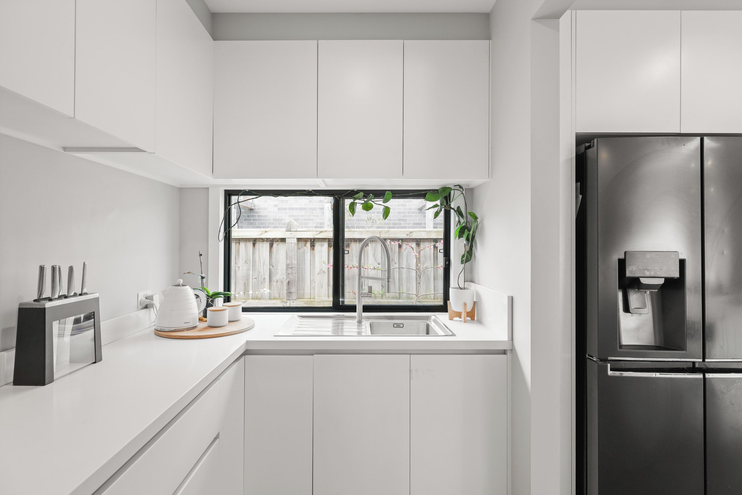 A modern kitchen with white cabinetry, a stainless steel refrigerator, a built-in coffee maker, and a window with a view of a wooden fence.