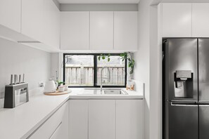 A modern kitchen with white cabinetry, a stainless steel refrigerator, a built-in coffee maker, and a window with a view of a wooden fence. - The Salty Snapper (Rhyll)
