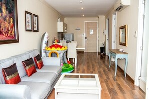 En stue med sofa, et lille bord, et farverigt legeområde for børn og et køkken i baggrunden.