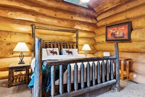 Ein Schlafzimmer in einer Blockhütte mit einem Himmelbett, zwei Nachttischlampen, einem Kissen mit Elchmotiv und einem gerahmten Bild von einem Elch an der Wand.