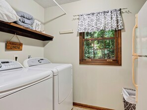 A laundry room with two front-loading washing machines, a wooden shelf with linens, a window with a view of greenery, and a sign indicating the room's purpose. - Cathedral Ceilings + Hot Tub + Game Room | Fairway Views | Creek & Green (Gatlinburg)