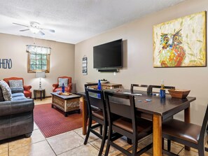 A living room with a sofa, a coffee table, a dining table with chairs, a television, and a painting on the wall. - Cathedral Ceilings + Hot Tub + Game Room | Fairway Views | Creek & Green (Gatlinburg)