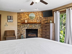 Ein Schlafzimmer mit Kamin aus Stein, einem Bett, einem Sessel, einem Fernseher und einem Fenster mit Vorhängen.
