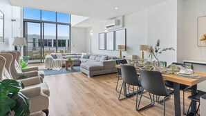 A modern living room with a dining area, large windows, and a view of the building outside. - Mascot Luxe 2BR | Airport | Pool + 2 Parking (Mascot)