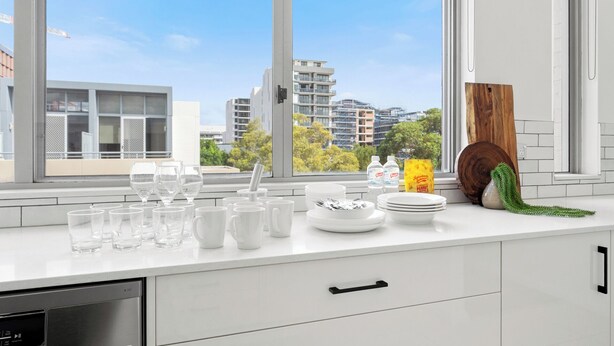 A modern kitchen with a large window, white cabinetry, and a view of buildings and trees. - Mascot Luxe 2BR | Airport | Pool + 2 Parking (Mascot)