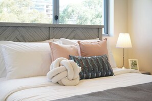 Una cama con sábanas blancas, un cojín decorativo, un suave plaid blanco y una lámpara de noche.