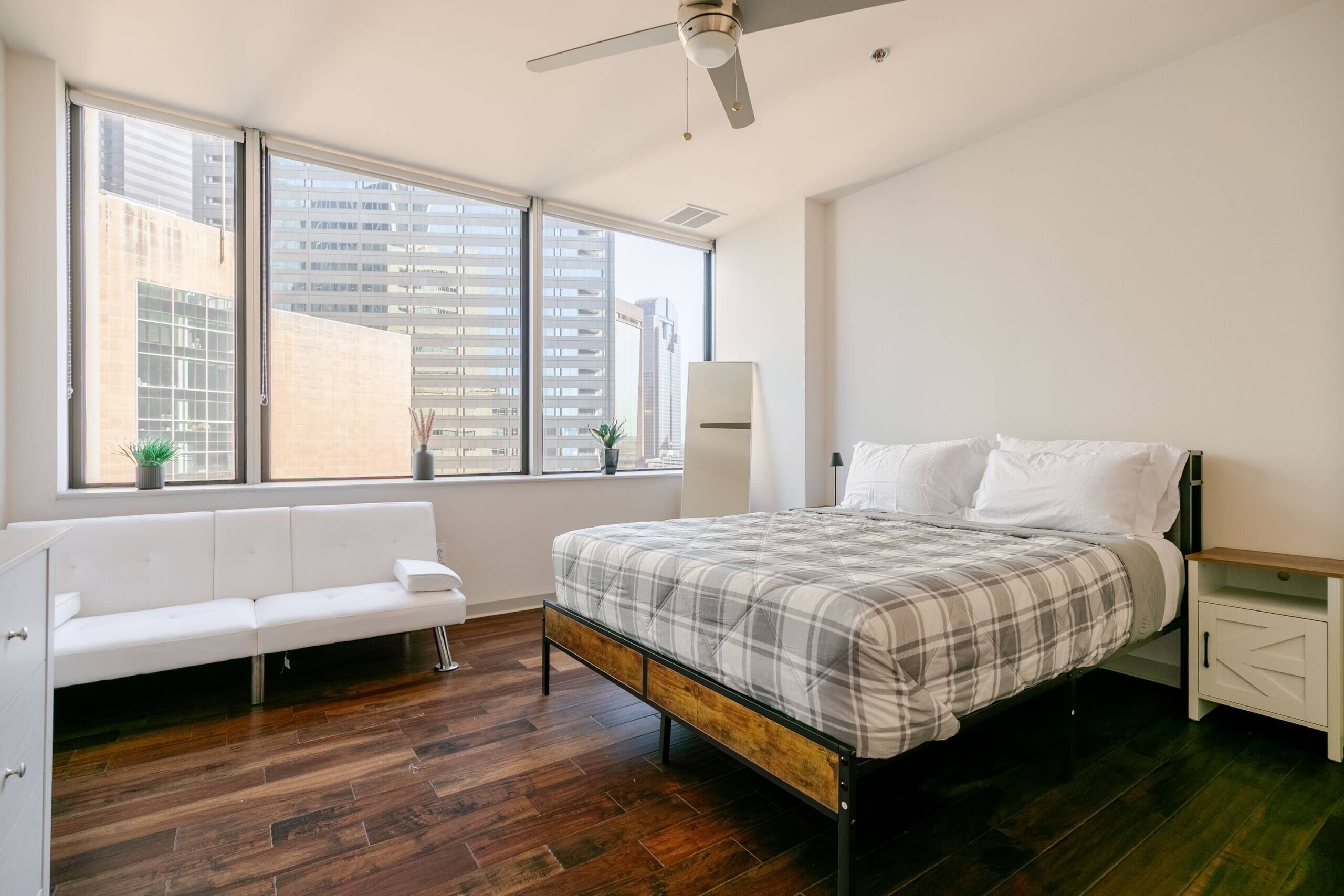 A bedroom with a bed, a white sofa, a nightstand, a lamp, a ceiling fan, and large windows with a city view.