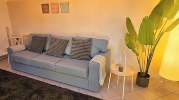 A living room with a light blue sofa, patterned cushions, a small side table with a candle, and a large green plant.