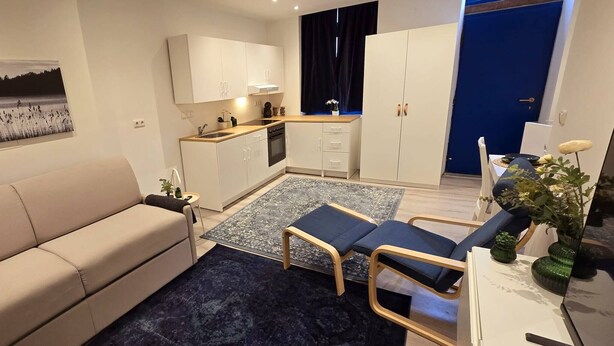 Un espace de vie compact comprenant un canapé, une petite table à manger, un coin cuisine et un rideau bleu.