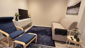 Un salon moderne avec un fauteuil bleu, un canapé beige, une petite table d’appoint, un téléviseur à écran plat et un tableau encadré accroché au mur.