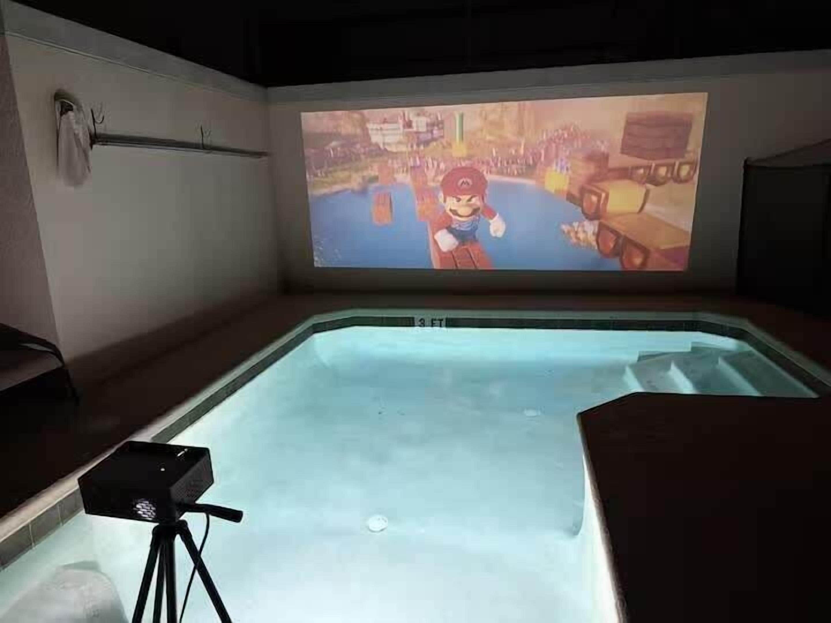 A room with a large screen displaying a video game, a pool, and a projector setup.