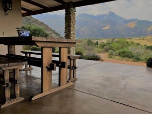 Een terras buiten met een stenen pilaar, een houten tafel en bankjes, met uitzicht op een bergachtig landschap.
