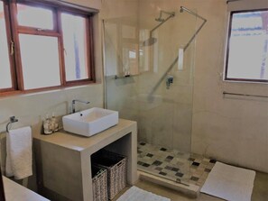 Een badkamer met een douchecabine van glas, een wit wastafeltje en een raam boven de spiegel.