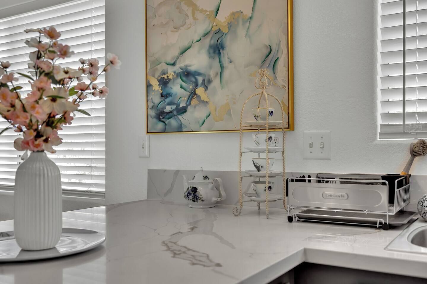 Une cuisine avec un plan de travail en marbre, un vase blanc rempli de fleurs, une théière, un service à thé et un tableau accroché au mur.