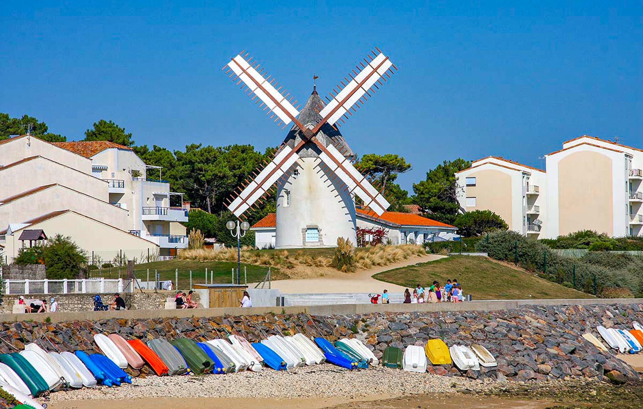 A windmill with a white body and red accents, surrounded by modern buildings and a beach with colorful kayaks.
