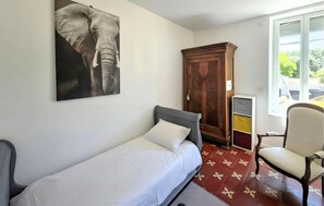 A single bed with a grey headboard, a wooden wardrobe, a chair, and a window with a view of greenery. - Charmante maison vendéenne (Longeville-sur-Mer)