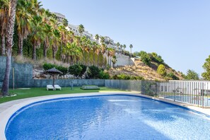 Ein Swimmingpool mit klarer, blauer Wasseroberfläche, umgeben von einer niedrigen Steinmauer und einer Rasenfläche mit zwei weißen Liegestühlen.