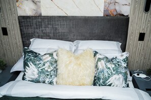 Ein Bett mit grauem Kopfteil, Kissen mit weiß-grünem Muster und einem flauschigen weißen Zierkissen.