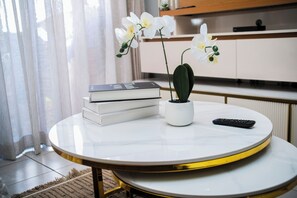 Ein runder Tisch mit weißer Oberfläche und goldener Kante, eine Topfpflanze, ein Bücherstapel und eine Fernbedienung.