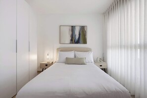A hotel room with a large bed, a bedside table, a nightstand, a wall-mounted artwork, and sheer curtains. - Cozy & Spacious 3BR  with Balcony & Mamad (Bat Yam)
