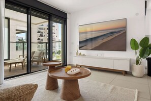 A modern living room with a large window, a wooden coffee table, and a painting of a beach scene. - Cozy & Spacious 3BR  with Balcony & Mamad (Bat Yam)