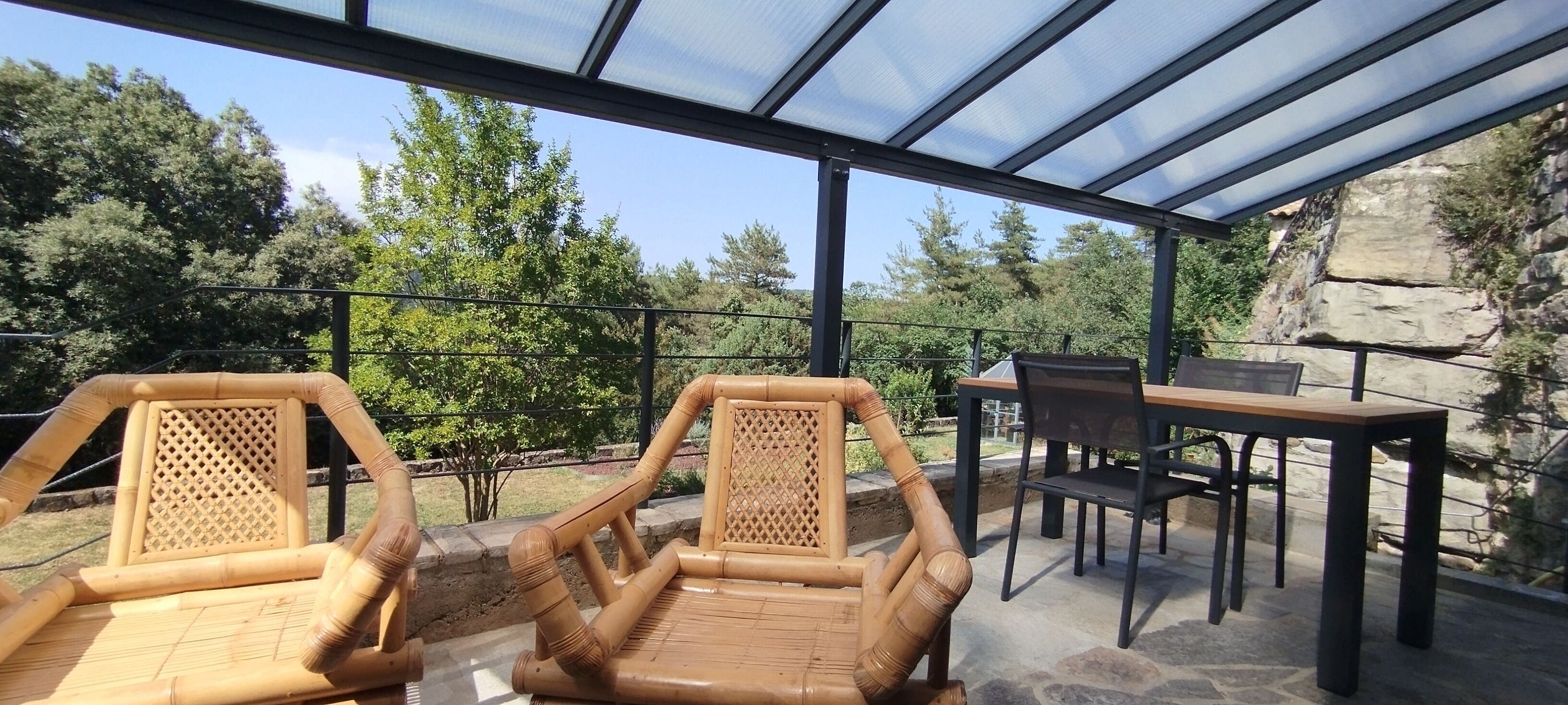 A covered outdoor seating area with two wicker chairs and a table, overlooking a rocky landscape and trees.