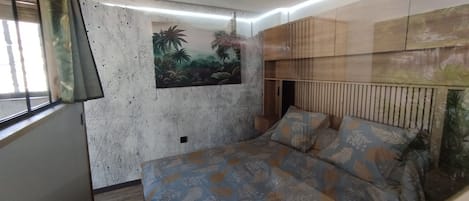 A room with a bed, a wall-mounted shelf, and a window with curtains.