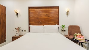 A hotel room with a wooden headboard, two bedside lamps, a bed with white linens, a brown chair, a small table with snacks, and two potted plants.
