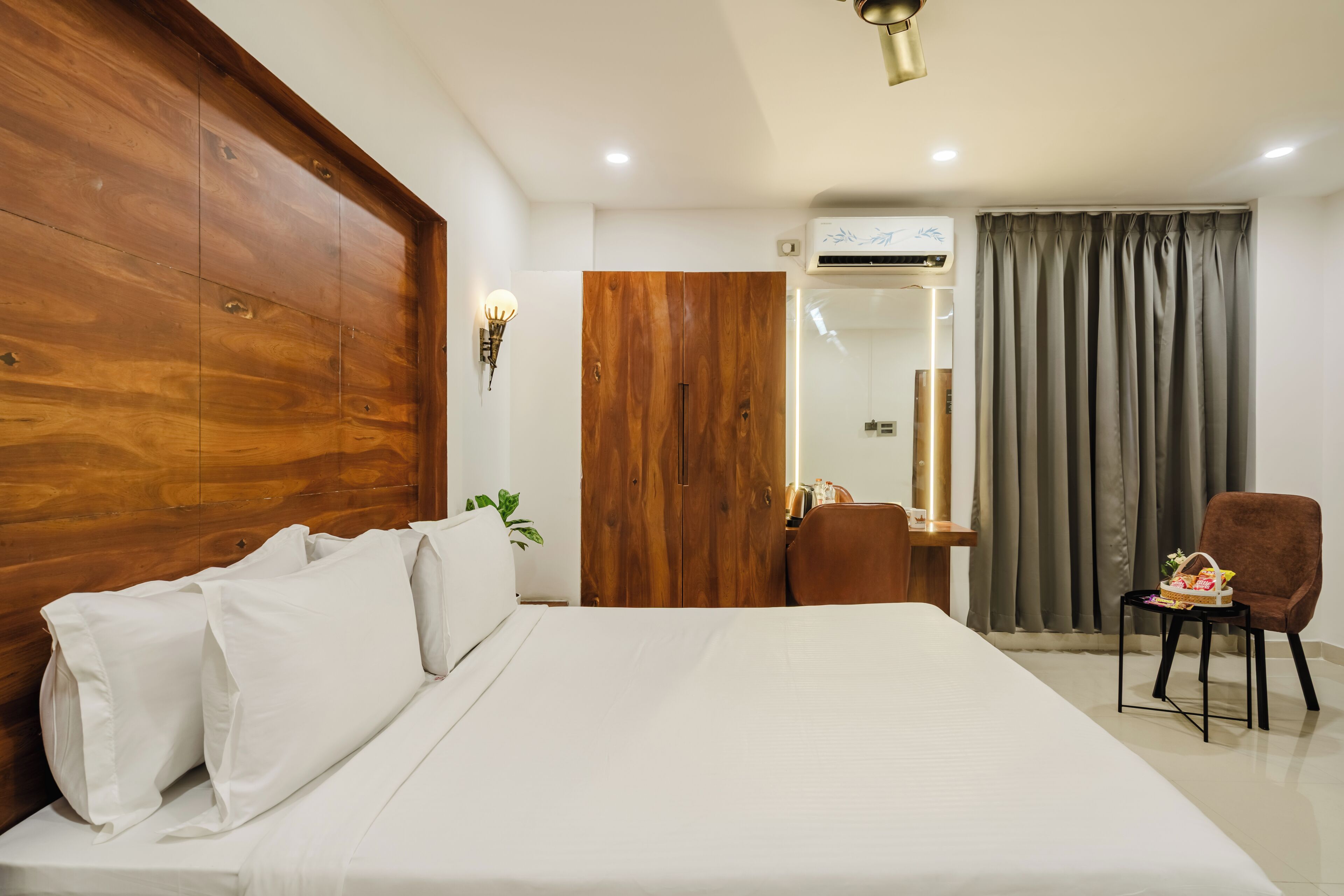 Une chambre d’hôtel avec un grand lit, une armoire en bois, un bureau avec une chaise, un miroir et une fenêtre avec des rideaux.