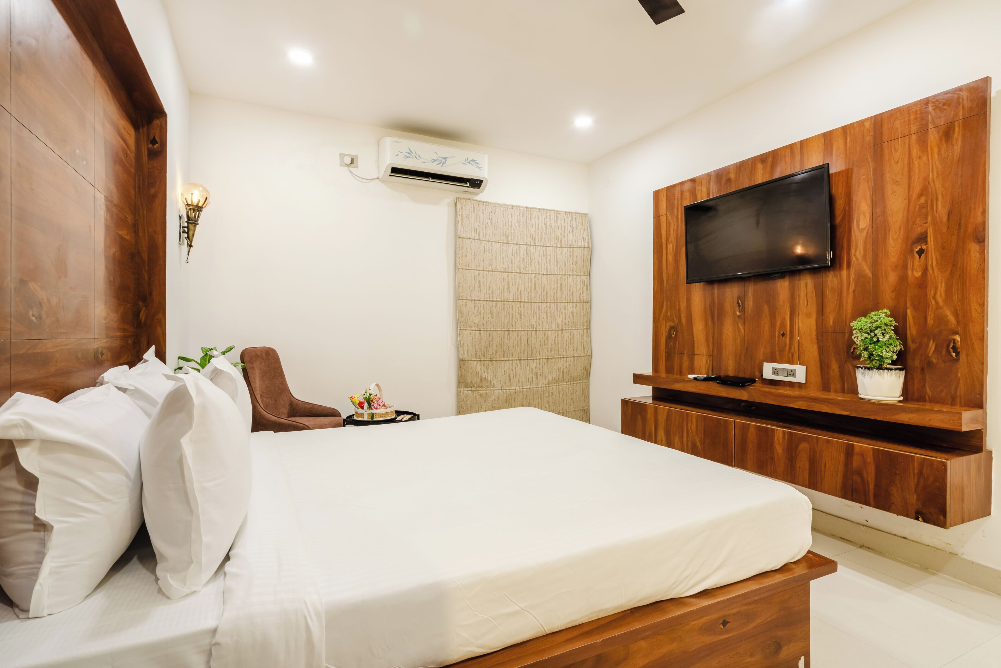 A bedroom with a bed, a TV mounted on a wooden wall, an air conditioning unit, and a small plant on a shelf.