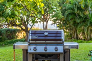 A Weber Genesis II gas grill with four burners, located outdoors with a garden in the background. - Spacious Ground-Floor 2BR with AC & Garden Views, Walk to Beach- Maui Kamaole F107 (Kihei)