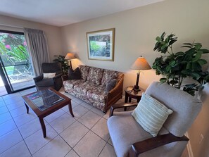 A living room with a sofa, armchair, coffee table, and lamps. There is a window with curtains, a framed picture on the wall, and a potted plant. - Spacious Ground-Floor 2BR with AC & Garden Views, Walk to Beach- Maui Kamaole F107 (Kihei)