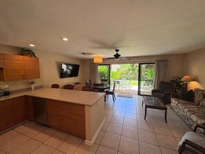 A spacious kitchen with a large island, dining area with chairs, a sofa, and a television mounted on the wall. - Spacious Ground-Floor 2BR with AC & Garden Views, Walk to Beach- Maui Kamaole F107 (Kihei)