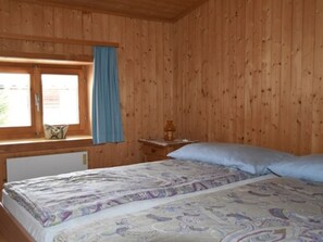 Una habitación de madera con una cama, un ventanal con cortinas y una mesita de noche con lámpara.