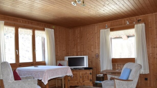 Interior de madeira com uma mesa de jantar, duas cadeiras, uma televisão e janelas com cortinas.