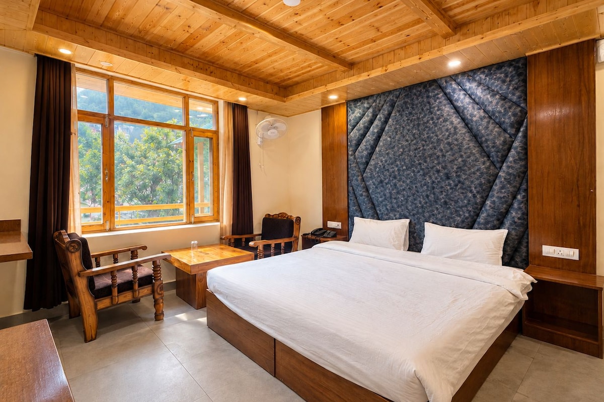 Une chambre d’hôtel avec un grand lit, des meubles en bois, un bureau et une vue sur la verdure par la fenêtre.