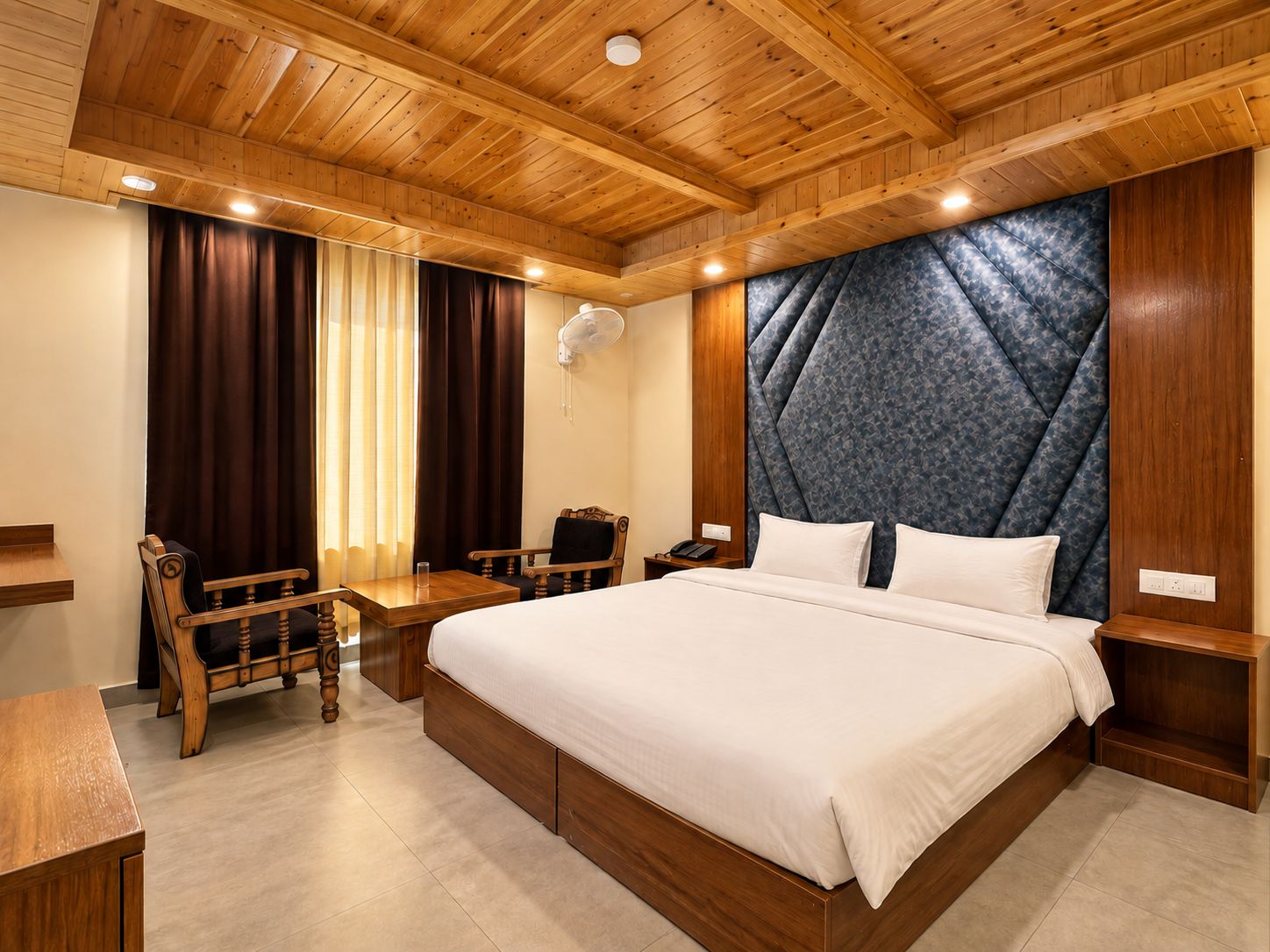 Una camera d'albergo con soffitto in legno, un letto grande con lenzuola bianche, un comodino in legno, una sedia di legno e una scrivania con una poltrona.